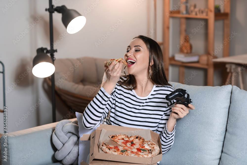 Beautiful young woman eating tasty pizza while playing video game at home in evening