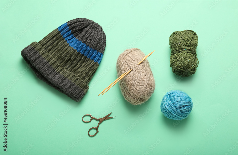 Yarn with knitting needles, hat and scissors on color background