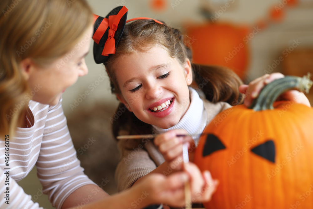 Happy mother and daughter making jack-o-lantern together in kitchen, drawing scary face on pumpkin