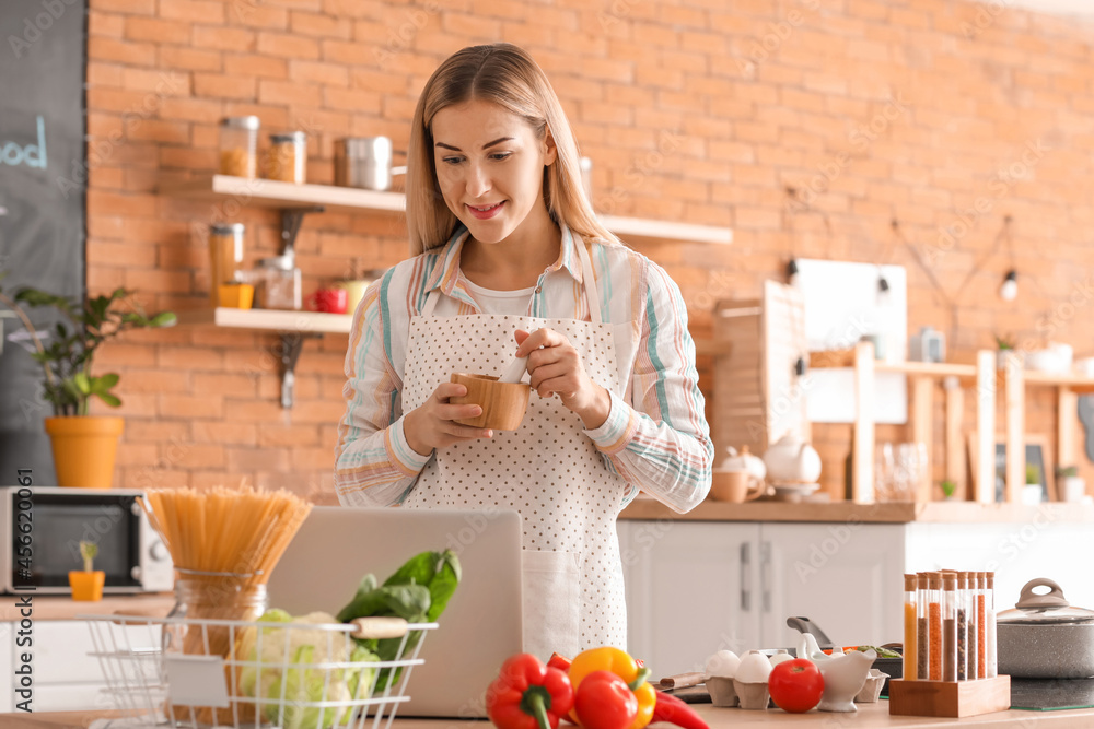 Young housewife with laptop cooking in kitchen at home