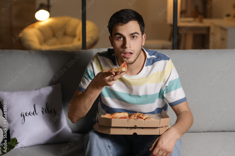 Handsome young man eating tasty pizza at home in evening