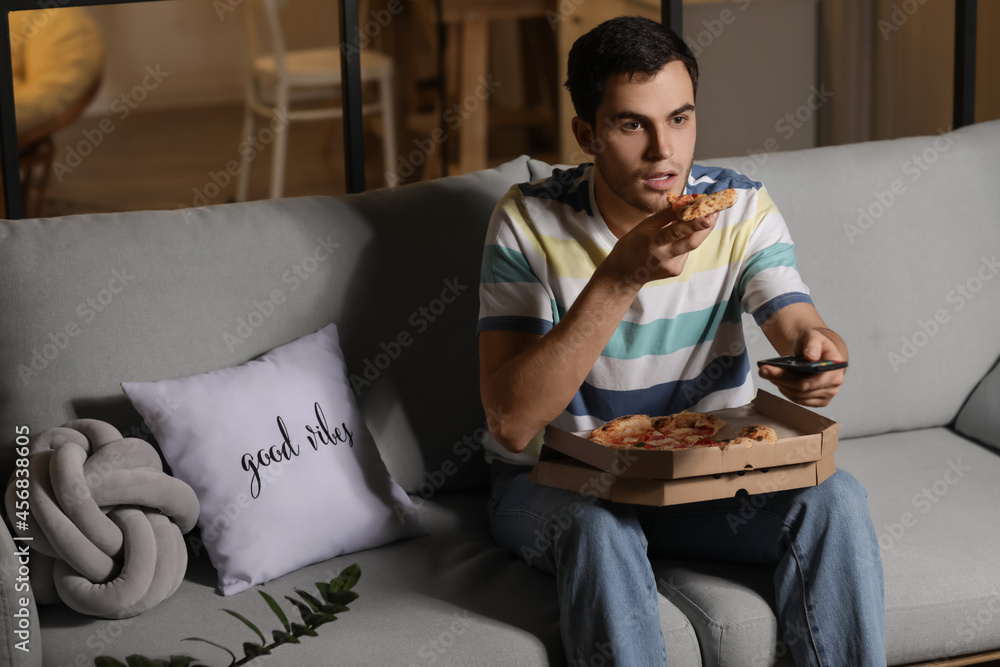 Handsome young man eating tasty pizza while watching TV at home in evening