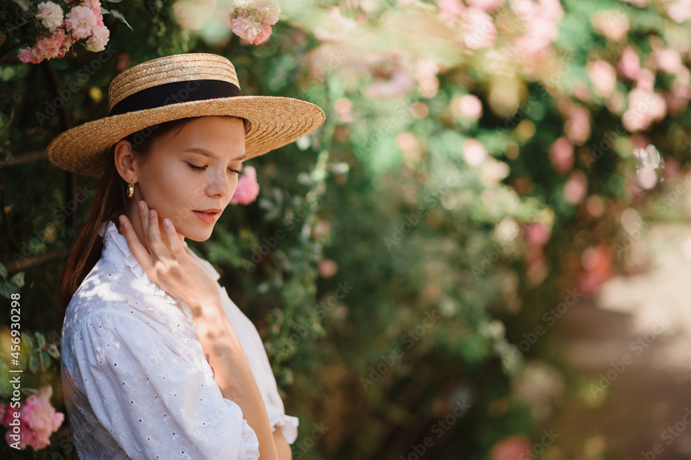 Elegant young woman wearing summer straw hat, white blouse, posing in blooming rose garden. Copy, em