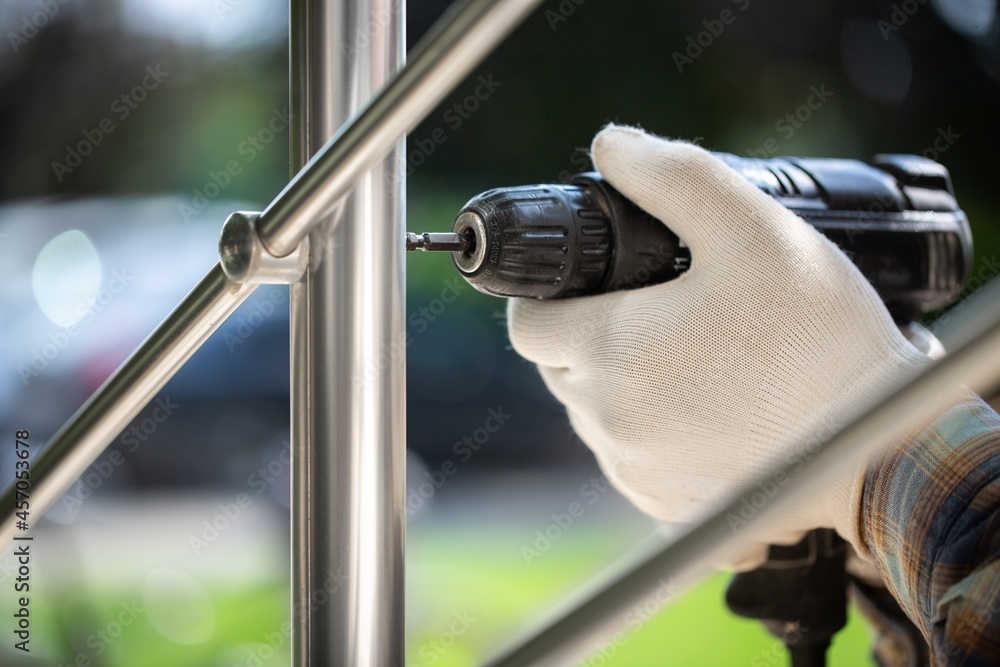 A worker in gloves repairs a metal railing of a staircase using a screwdriver and a drill