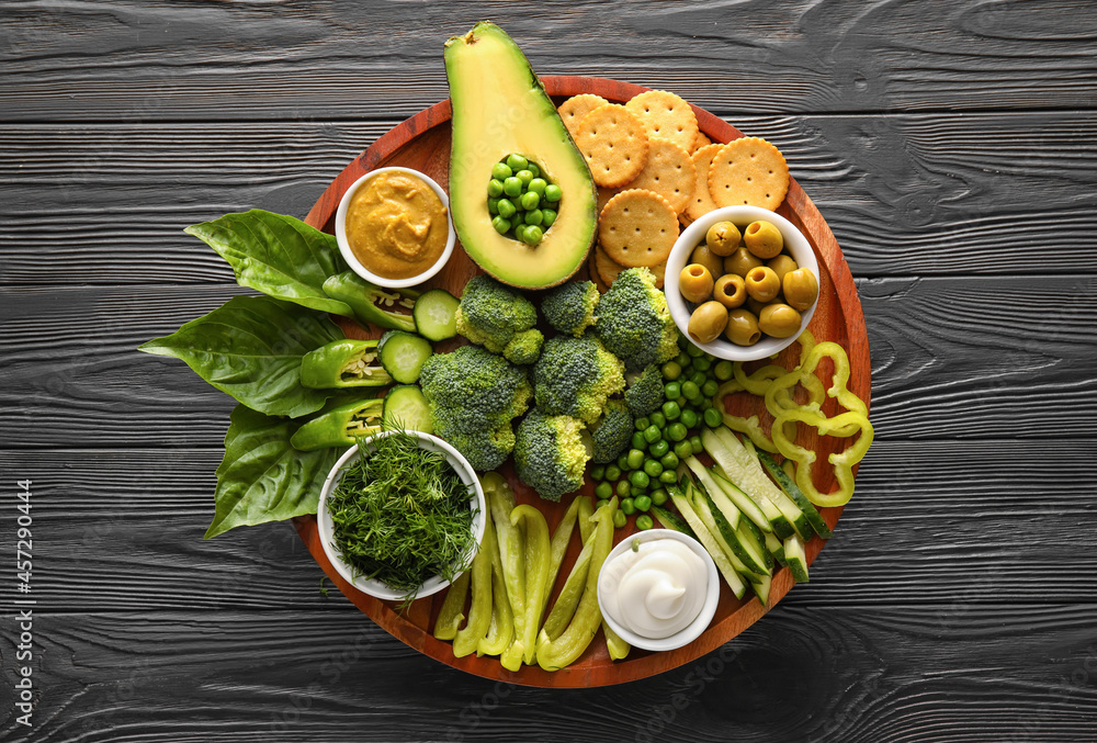 Stand with different green vegetables and bowls with sauce on dark wooden background