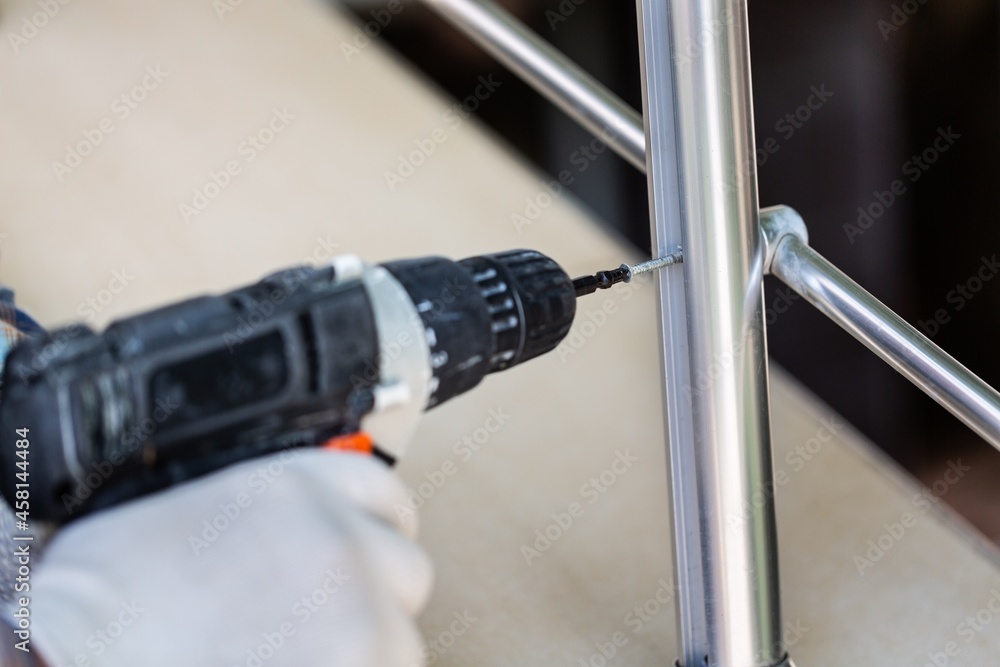 A worker in gloves repairs a metal railing of a staircase using a screwdriver and a drill