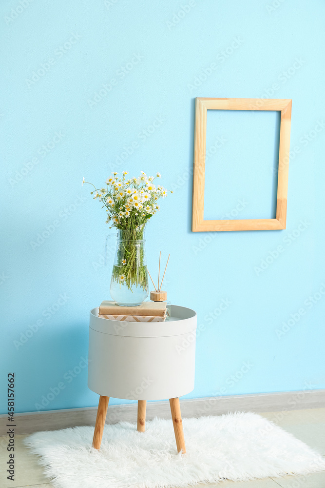 Vase with chamomiles, books and reed diffuser on table near color wall