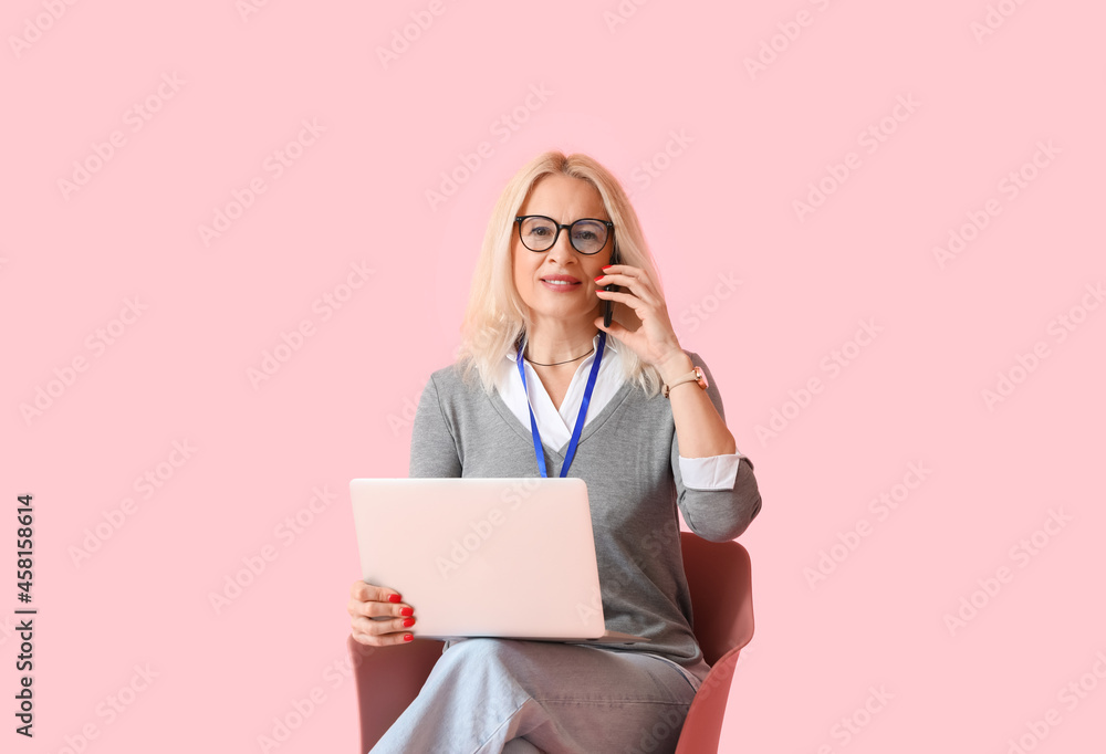 Portrait of mature businesswoman with laptop talking by phone while sitting on chair against color b