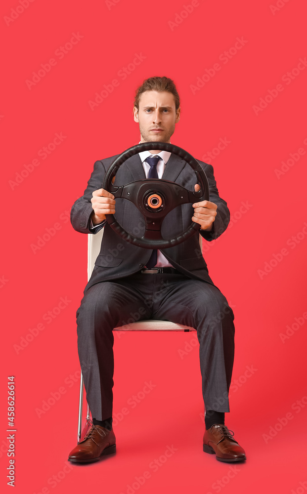 Young businessman with steering wheel sitting on chair against color background