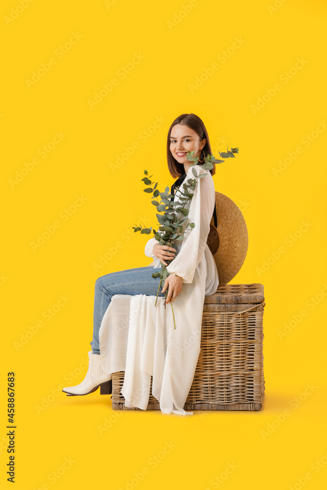 Beautiful young woman with eucalyptus branches sitting on wicker box against color background