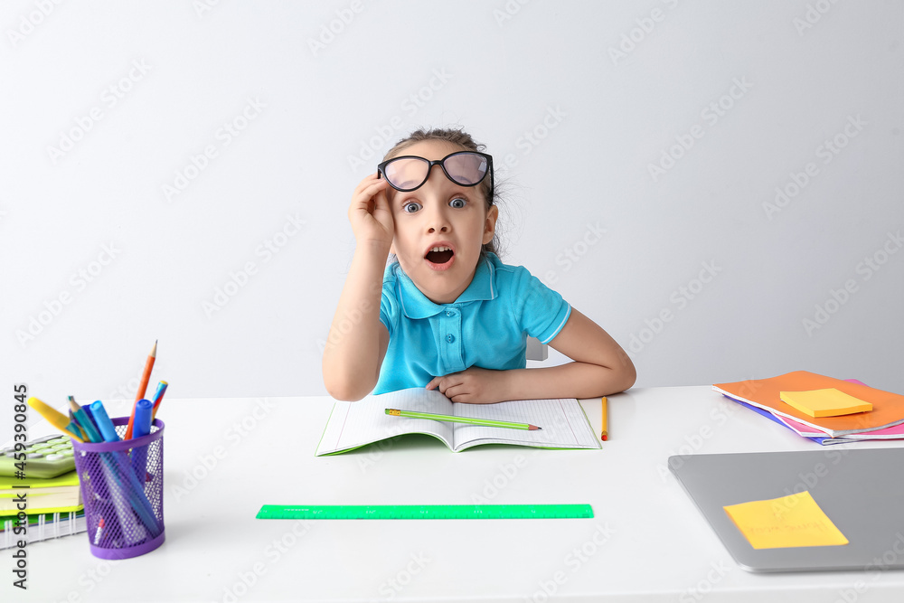 Shocked little girl doing homework at table on light background