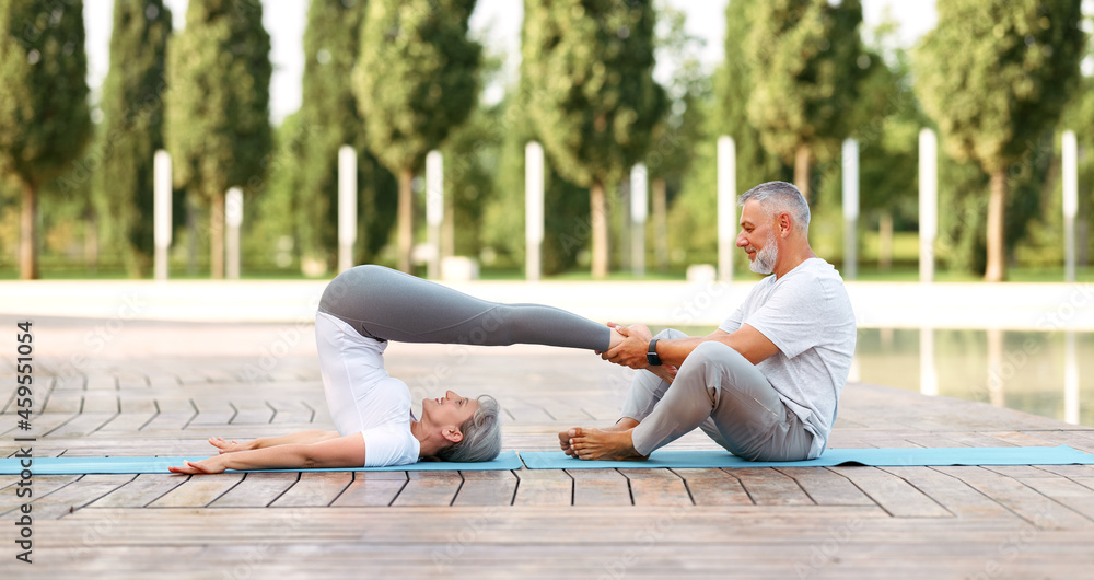 Elderly couple of husband and wife performing partner yoga, practicing Halasana on fresh air outside