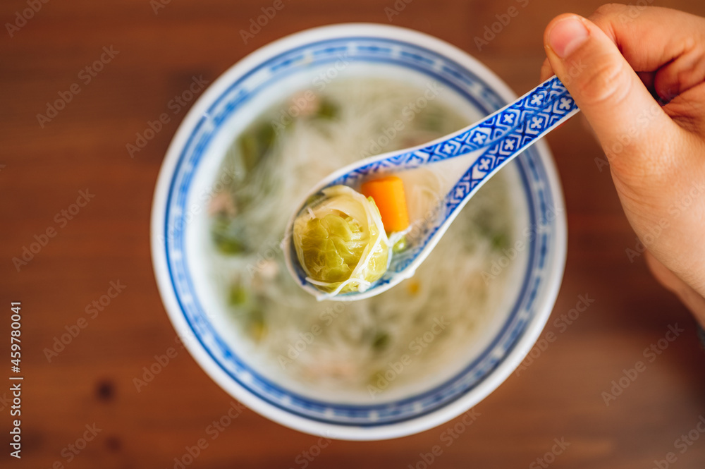 Top down view of a bowl of soup and a hand holding a spoon with cabbage and carrots and noodles.