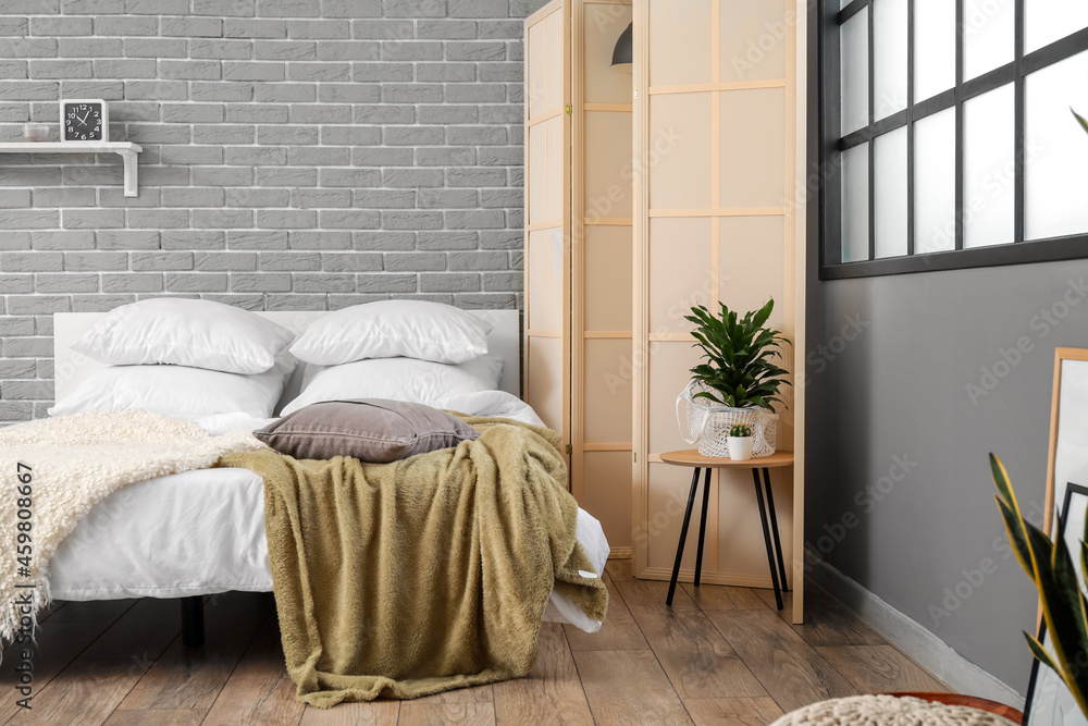 Comfortable bed, folding screen and table with houseplant near grey brick wall