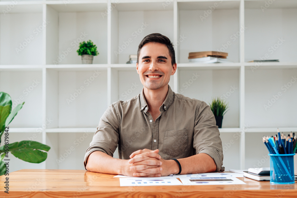 Portrait of happy businessman sitting at office desk, looking at camera, smiling.