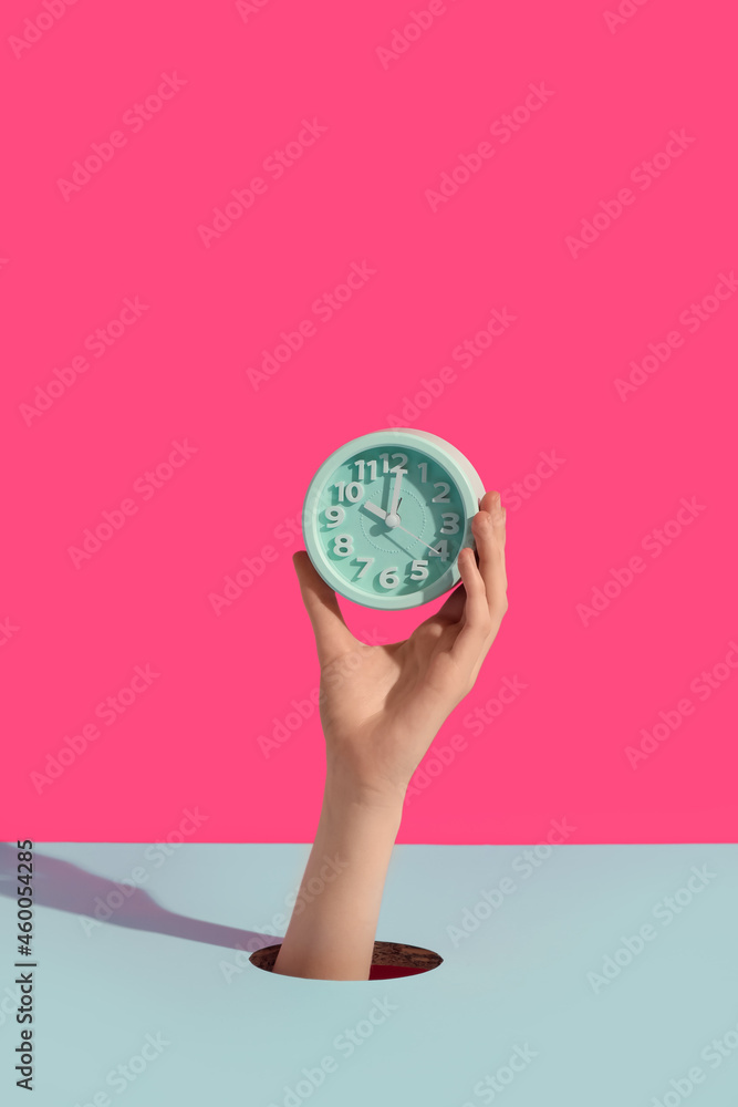 Female hands holding alarm clock through hole on color background