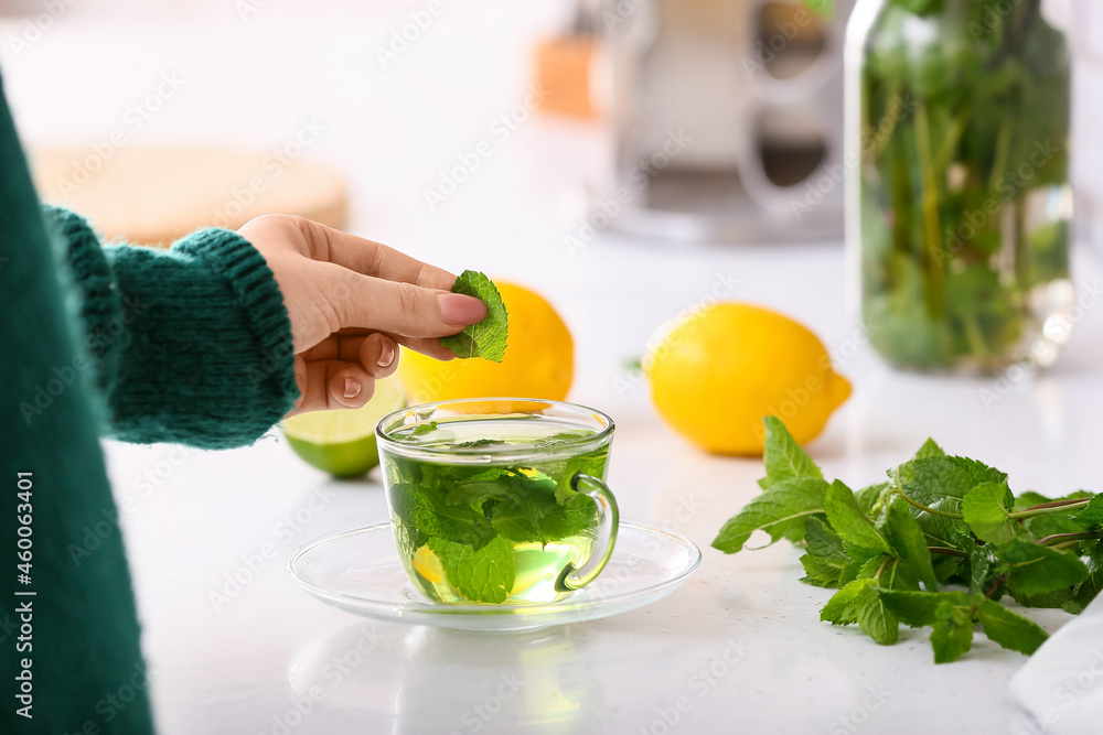 Woman putting mint leaf into glass cup with tea in kitchen, closeup