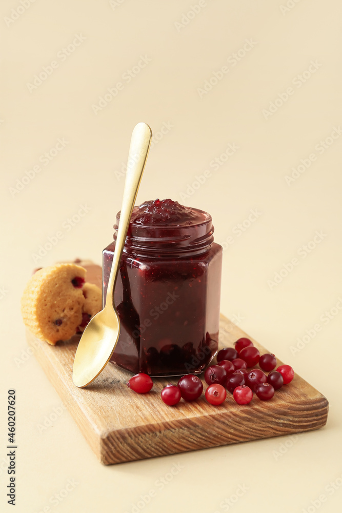 Glass jar of tasty cranberry jam and muffin on color background