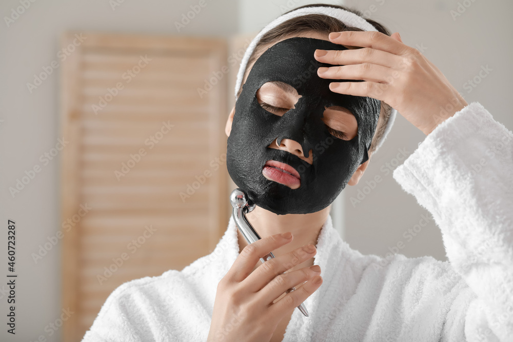 Beautiful young woman with sheet facial mask and massage tool at home