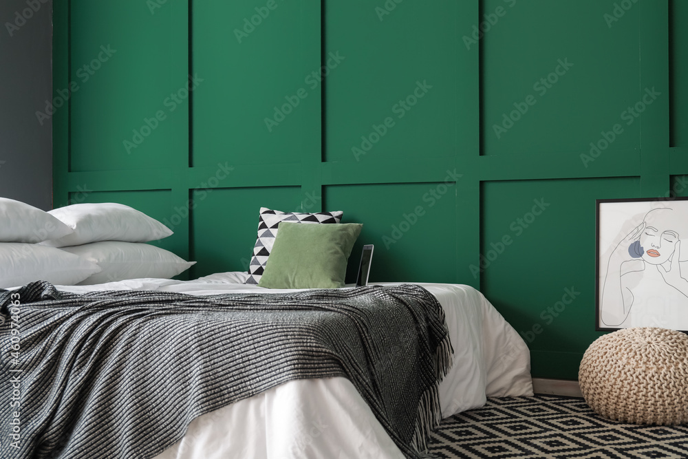 Comfortable bed with laptop, pillows and ottoman near green wall