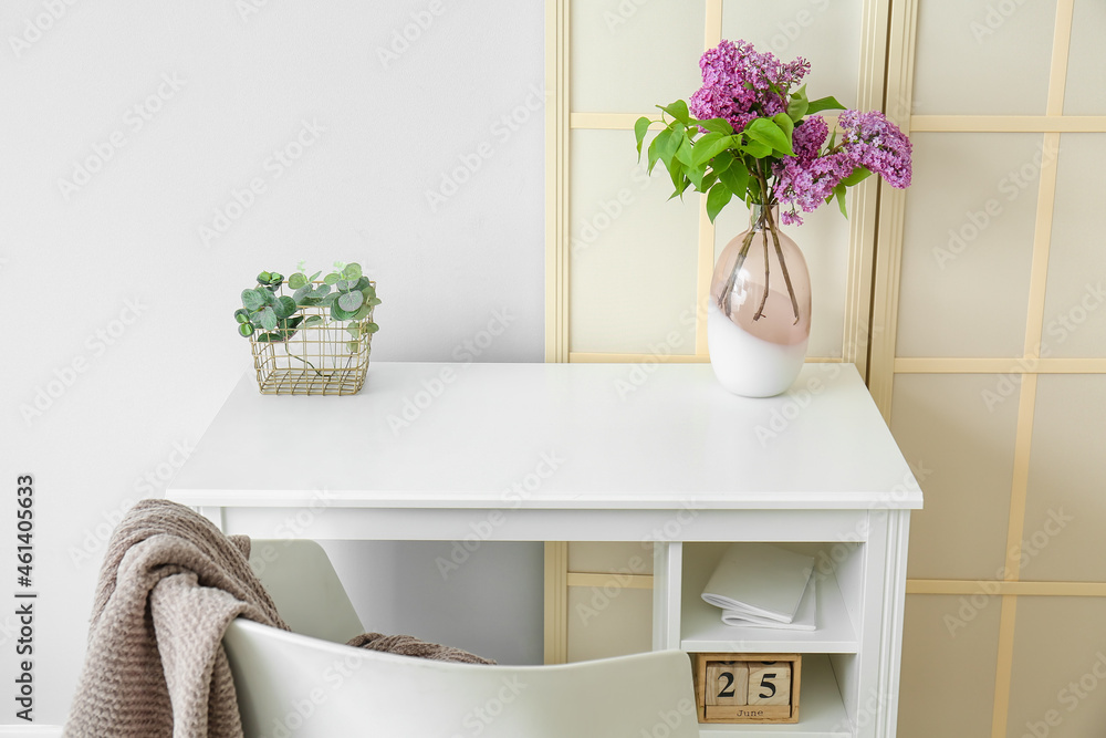 Desk with lilac flowers in vase and folding screen near light wall