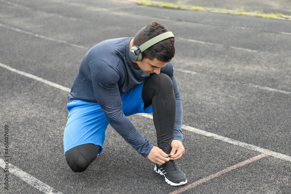 Muscular young man in headphones tying shoe lace at stadium