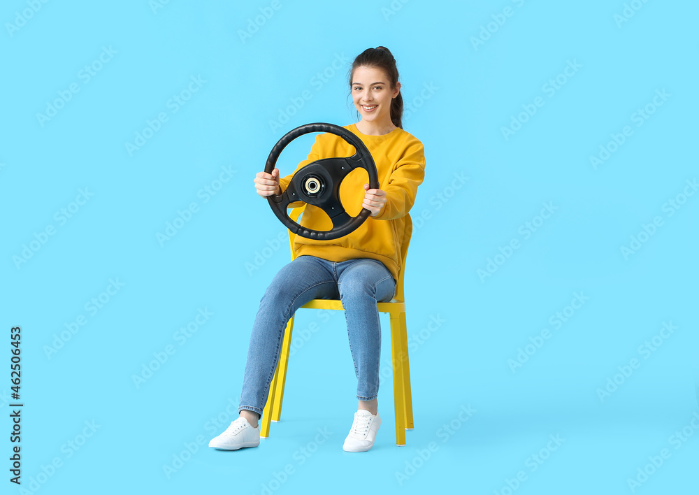 Young woman with steering wheel sitting in chair on color background