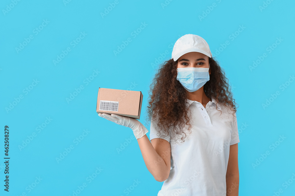 Female African-American courier in medical mask and with parcel on color background