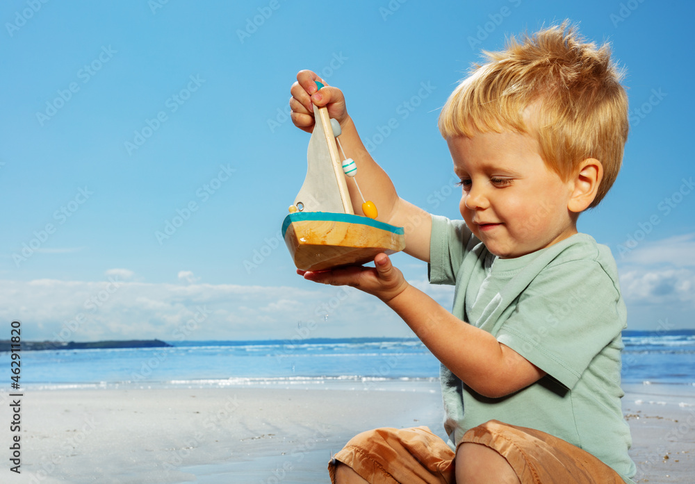 Cute blond boy play with sail boat yacht on beach