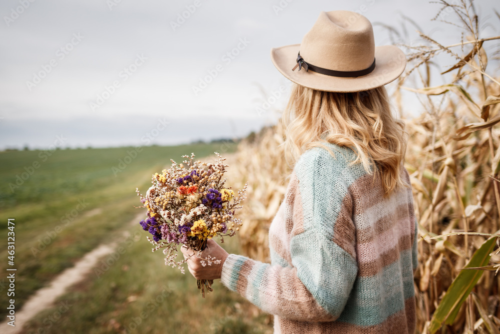 Woman with knitted sweater and hat holding bouquet of dried flowers outdoors. Herbs and blossom drie