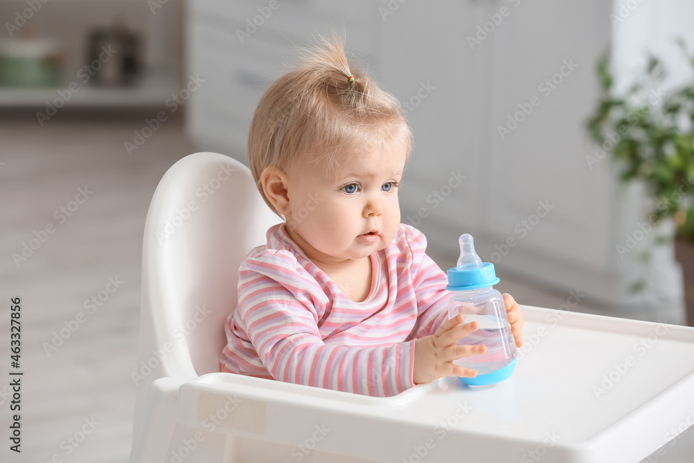 Cute baby girl with bottle of water sitting on high chair in kitchen