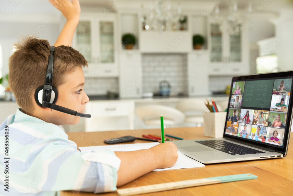 Caucasian boy raising hand for video call, with smiling diverse elementary school pupils on screen