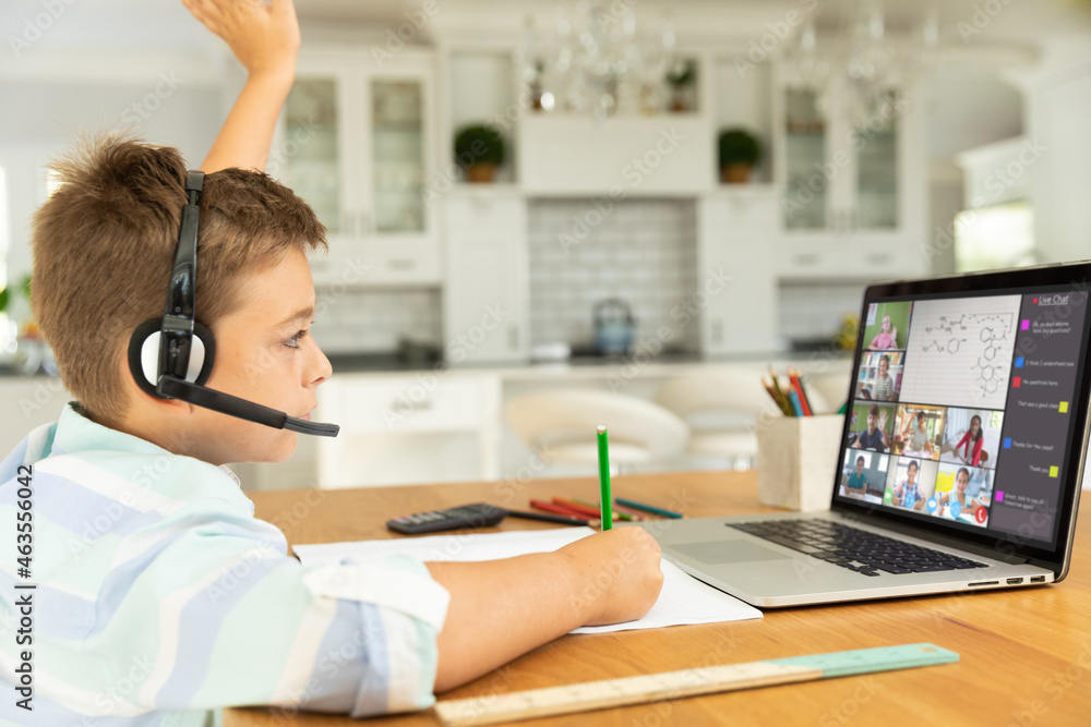 Caucasian boy raising hand for video call, with smiling diverse elementary school pupils on screen