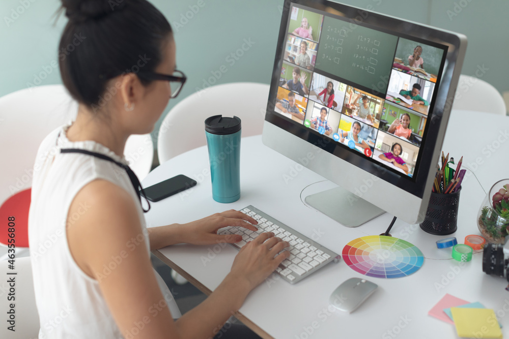 Asian girl using computer for video call, with smiling diverse elementary school pupils on screen