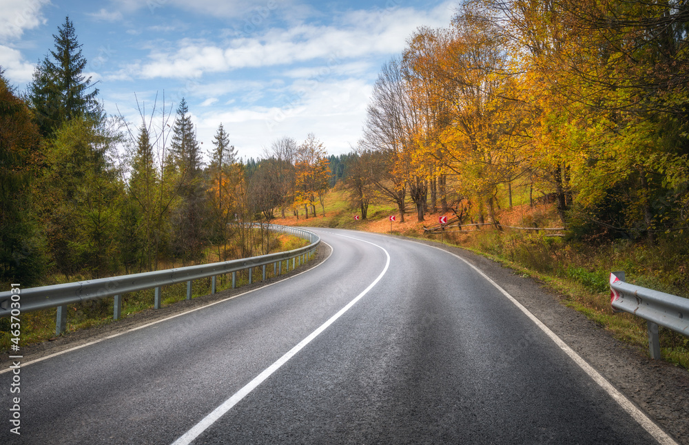 Road in autumn forest. Beautiful empty mountain roadway, trees with orange foliage and overcast sky.