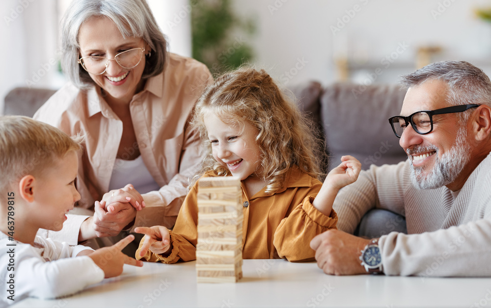 Excited children playing game Jenga at home with positive senior grandparents while sitting on sofa