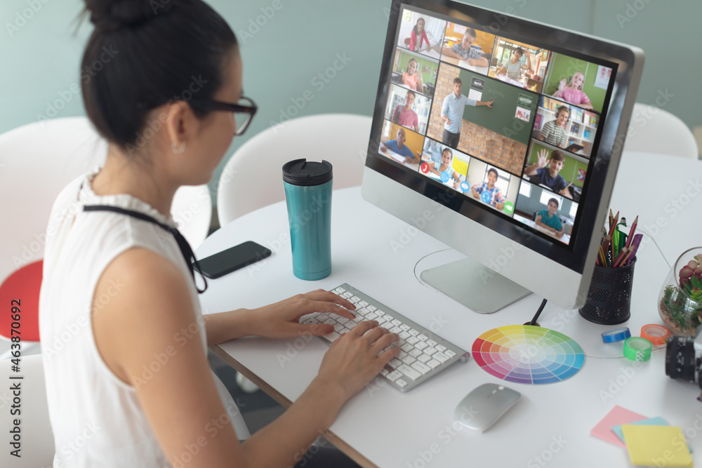 Asian girl using computer for video call, with smiling diverse elementary school pupils on screen