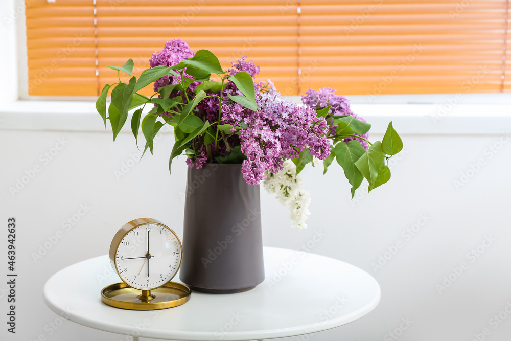 Clock and glass with lilac flowers in interior of light living room