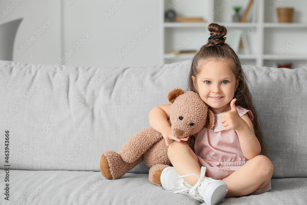 Adorable little girl with teddy bear showing thumb-up at home