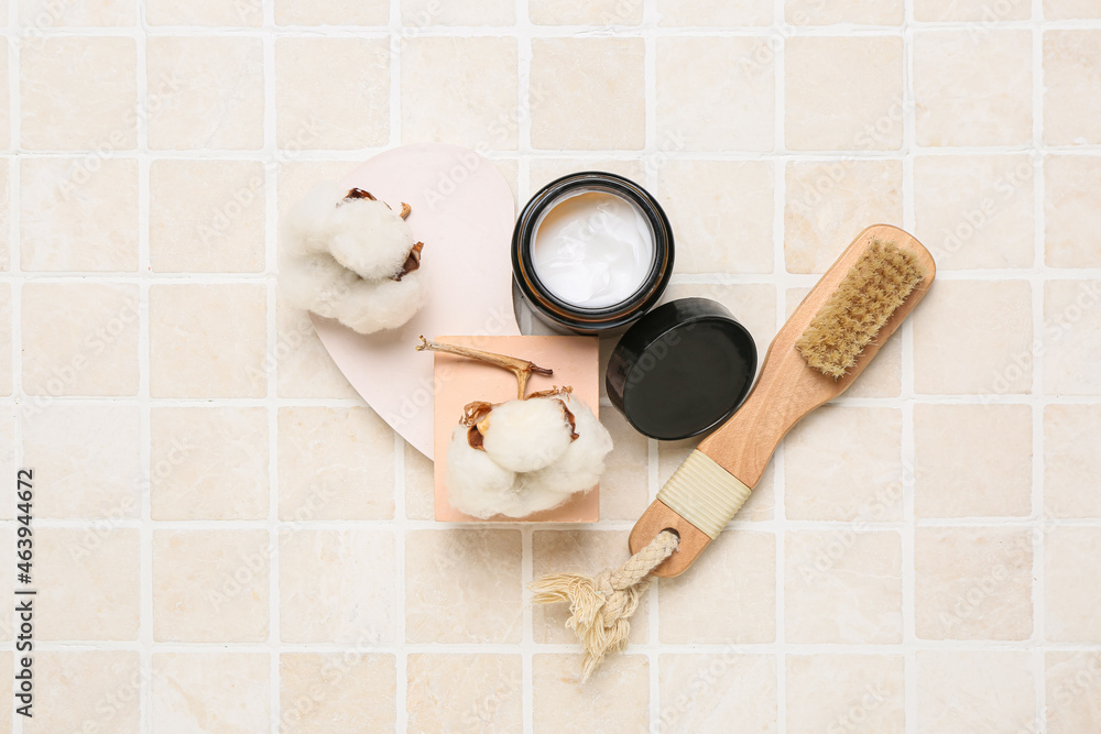 Composition with jar of cream, massage brush and cotton flowers on light tile