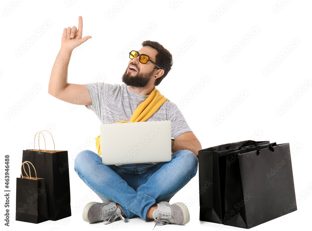 Handsome man with laptop and Black Friday shopping bags pointing at something on white background