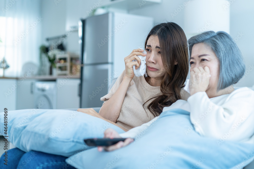 亚洲资深成熟女性和女儿坐在沙发上看悲伤的电影。