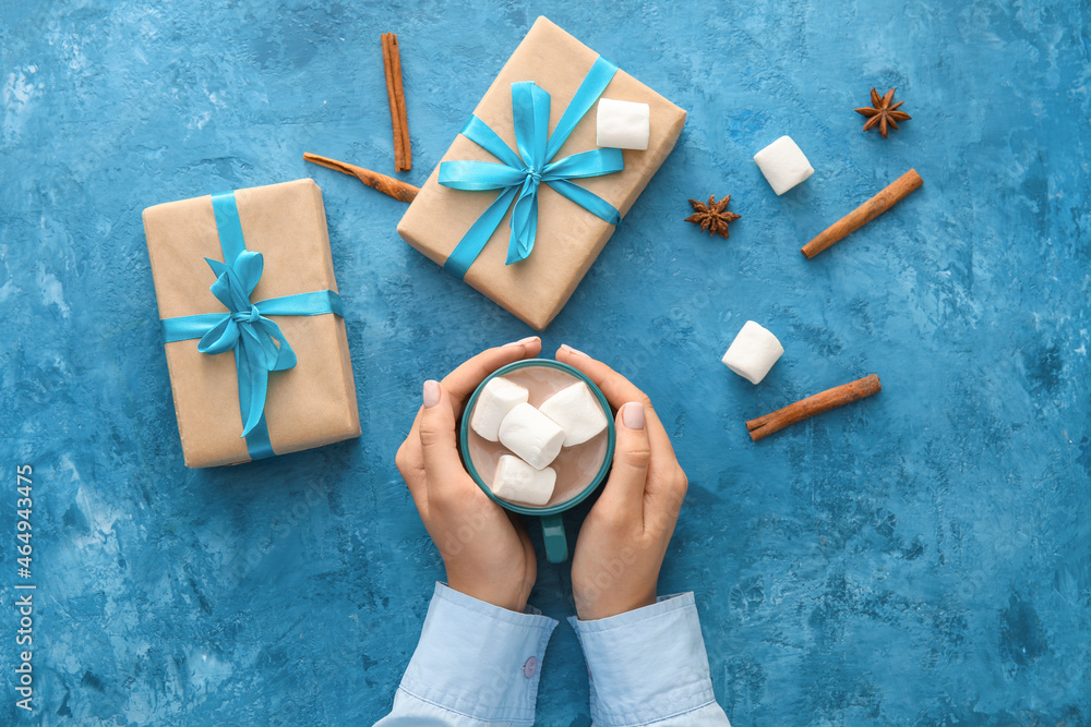 Female hands with cup of cacao and gifts on color background