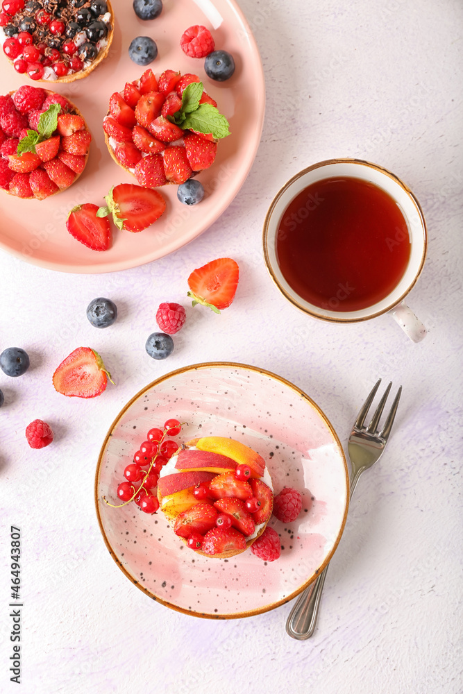 Plates with tasty berry tartlets and cup of tea on light background