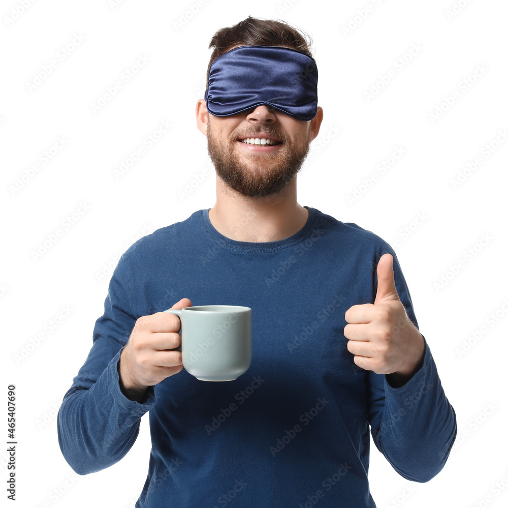 Young man in sleeping mask with cup of coffee showing thumb-up on white background