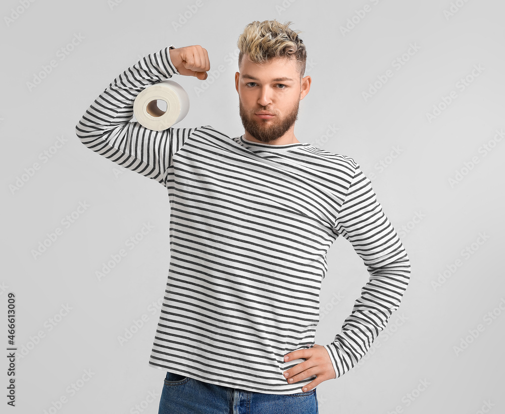 Muscled young man with toilet paper on light background