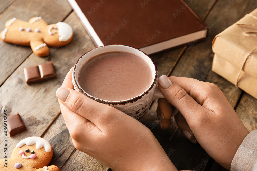 Female hands with cup of cacao, cookies, book and gift on wooden table, closeup