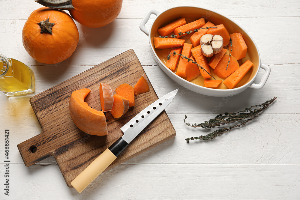 Baking dish and board with fresh pumpkin pieces on white wooden background