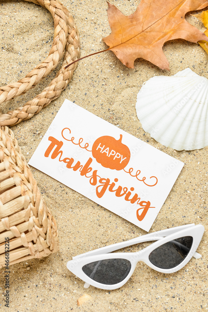 Wicker bag, dry leaf, seashell, sunglasses and paper card with text HAPPY THANKSGIVING on sand