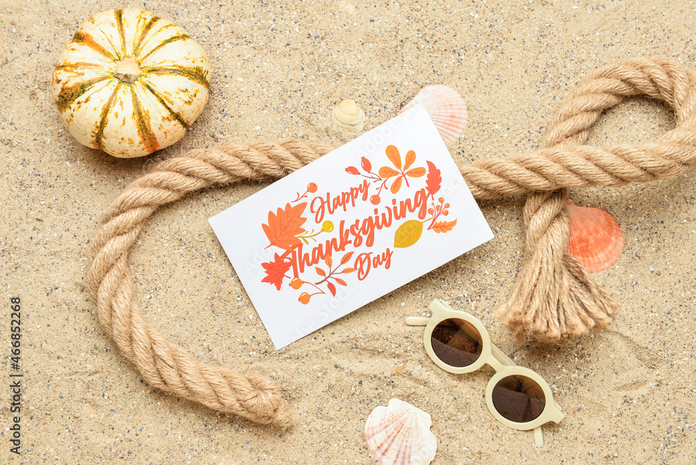 Pumpkin, rope, seashells, sunglasses and paper card with text HAPPY THANKSGIVING DAY on sand backgro